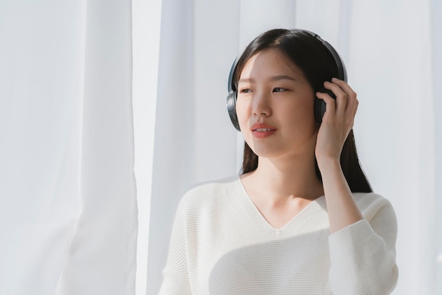 Relex y una mujer asiática pacífica disfrutan el fin de semana con la luz del sol de la mañana de los auriculares de música cerca del fondo de la casa de la ventana