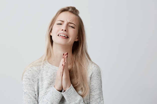 Religiosa supersticiosa rezando hermosa mujer con cabello rubio y liso, llorando, presionando las palmas juntas para tener buena suerte, esperando que los deseos se hagan realidad, con una mirada emocionada. Emociones humanas, sentimientos