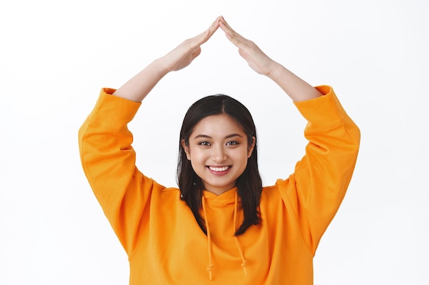 Relleno de niña asegurado. Retrato de primer plano linda mujer asiática tonta en sudadera con capucha naranja haciendo techo con las manos por encima de la cabeza sonriendo con alegría, concepto de seguridad