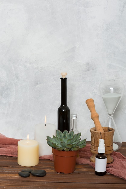 Reloj de arena planta de cactus; vela encendida el último; aceite esencial; Mortero de madera y pastel en mesa contra pared.
