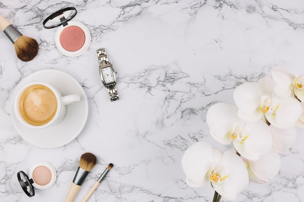 Reloj de pulsera; taza de café; polvo compacto; pincel de maquillaje y flor de la orquídea sobre fondo de mármol