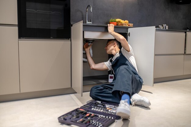 Reparación del hogar Hombre de mediana edad inspeccionando la tubería tocando la mano debajo del fregadero en una cocina moderna y elegante