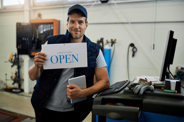 Reparador de automóviles feliz con cartel abierto y mirando a la cámara mientras está de pie en un taller