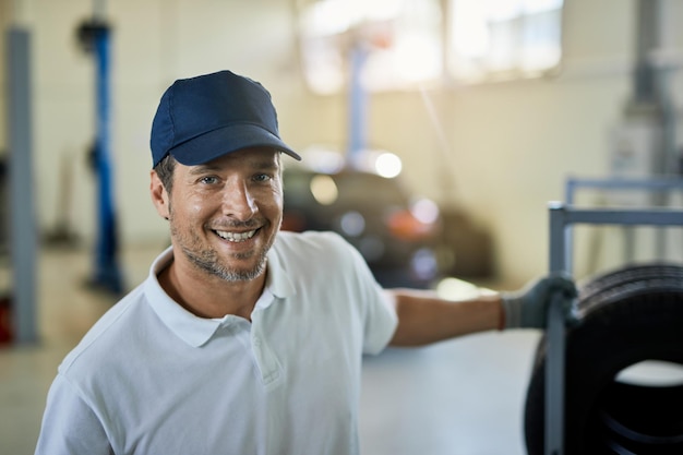 Reparador de automóviles feliz tirando de estante con neumáticos de coche y mirando a la cámara en un taller