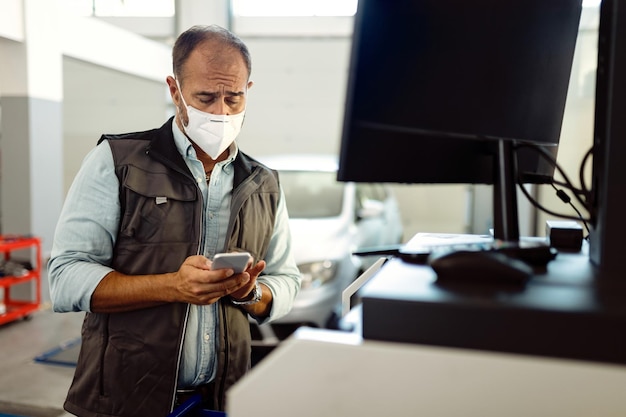 Reparador de automóviles con mascarilla protectora enviando mensajes de texto en un teléfono inteligente en su taller