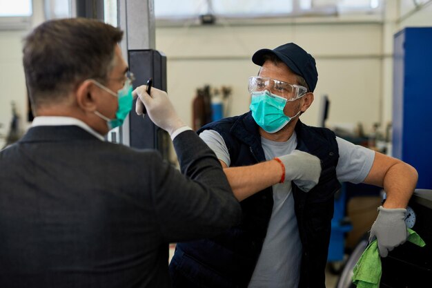 Reparador de automóviles y su gerente con máscaras protectoras y saludo con codos en un taller