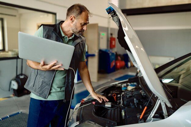 Reparador de automóviles usando una computadora portátil mientras examina el motor del automóvil en un taller