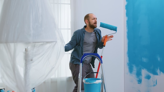 Reparador cantando sobre un cepillo de rodillo sumergido en pintura azul mientras redecora el apartamento. Tareas domésticas, diseño, renovación. Construcción de viviendas mientras se renueva y mejora. Reparación y decoración.