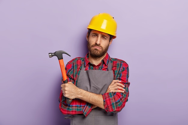 El reparador masculino infeliz sombrío tiene una mirada triste y cansada, mantiene las manos cruzadas, sostiene el martillo en la mano, se fatiga después de la reparación y el trabajo manual, usa un uniforme especial. Artesanía, martilleo, construcción.