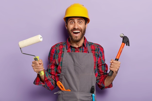 El reparador positivo sostiene un martillo y un rodillo de pintura, usa delantal y casco, tiene muchas herramientas de construcción, listas para renovar la casa. Feliz hombre de servicio profesional puede reparar todo en su apartamento