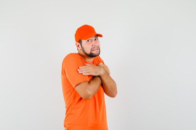 Repartidor abrazándose a sí mismo en camiseta naranja, gorra y mirando humilde, vista frontal.