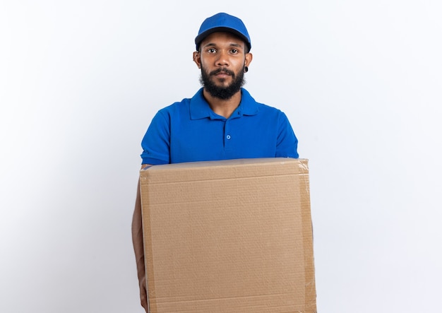 Repartidor afroamericano joven confiado que sostiene la caja de cartón grande aislada en la pared blanca con el espacio de la copia