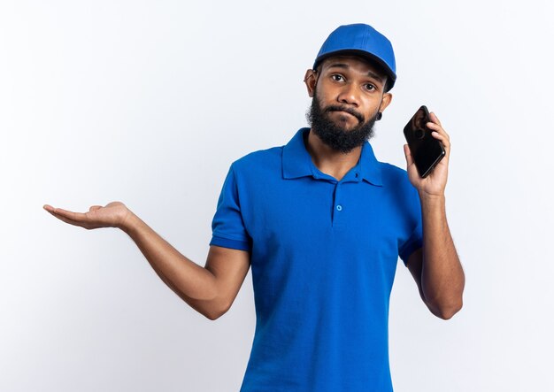 Repartidor afroamericano joven decepcionado hablando por teléfono y manteniendo la mano abierta aislada sobre fondo blanco con espacio de copia