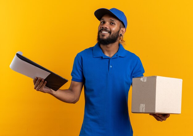 Repartidor afroamericano joven satisfecho con caja de cartón y portapapeles aislado en la pared naranja con espacio de copia