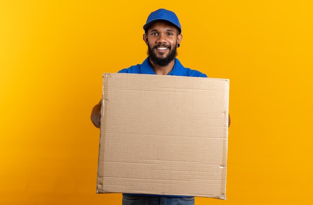 Repartidor afroamericano joven sonriente que sostiene una caja de cartón grande aislada en la pared naranja con espacio de copia