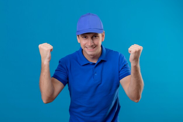 Repartidor apuesto joven en uniforme azul y gorra apretando los puños feliz y salido regocijándose de su éxito de pie sobre la pared azul