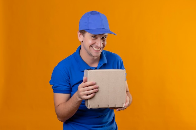 Repartidor apuesto joven en uniforme azul y gorra con paquete de caja sonriendo astutamente feliz y positivo de pie sobre la pared azul