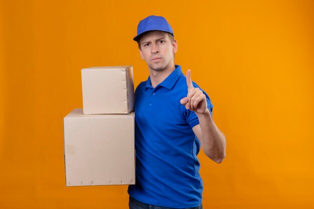 Repartidor apuesto joven en uniforme azul y gorra sosteniendo cajas de cartón señalando con el dedo hacia arriba advertencia con expresión seria en la cara de pie sobre la pared naranja