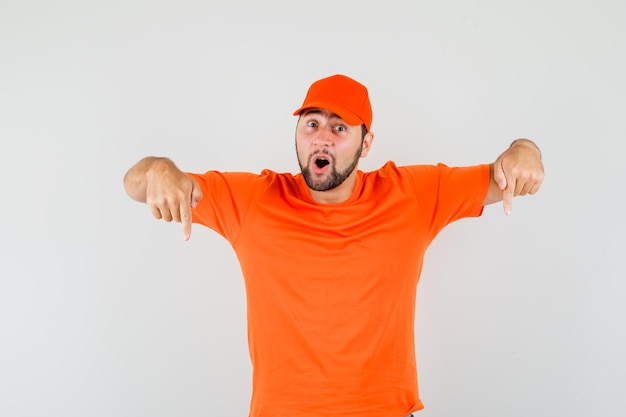 Repartidor apuntando hacia abajo en camiseta naranja, gorra y mirando asombrado, vista frontal.