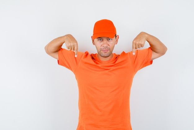 Foto gratuita repartidor apuntando hacia abajo en camiseta naranja y gorra y mirando seguro