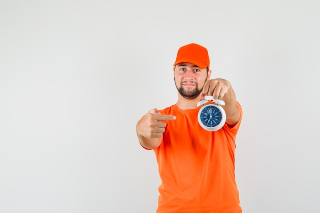 Repartidor apuntando al despertador en camiseta naranja, gorra y mirando confiado, vista frontal.