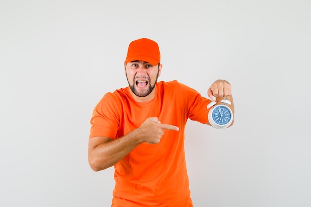 Repartidor apuntando al despertador mientras grita en camiseta naranja, gorra y mirando agitado, vista frontal.