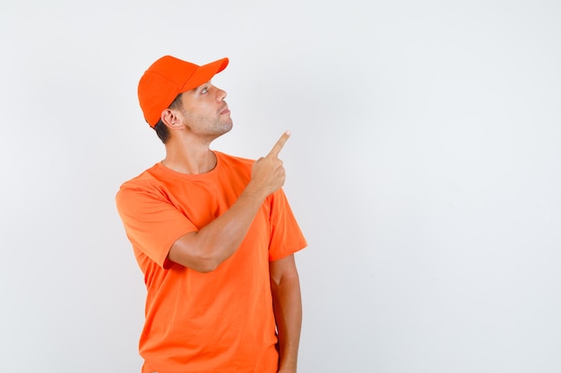 Foto gratuita repartidor apuntando hacia arriba mientras mira hacia arriba con camiseta naranja y gorra y mirando enfocado
