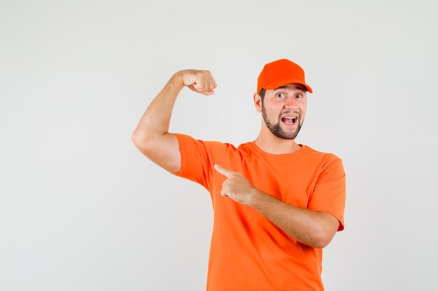 Repartidor apuntando a los músculos del brazo en camiseta naranja, gorra y mirando confiado, vista frontal.