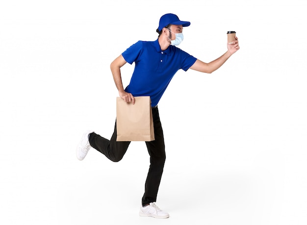 Repartidor asiático con mascarilla en uniforme azul corriendo con bolsa de papel y café para llevar aislado sobre un espacio en blanco. servicio de entrega urgente durante covid19.