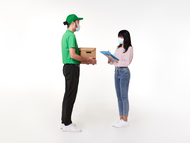 Repartidor asiático con mascarilla en uniforme verde con caja de paquetería para dar a la mujer cliente sobre blanco.