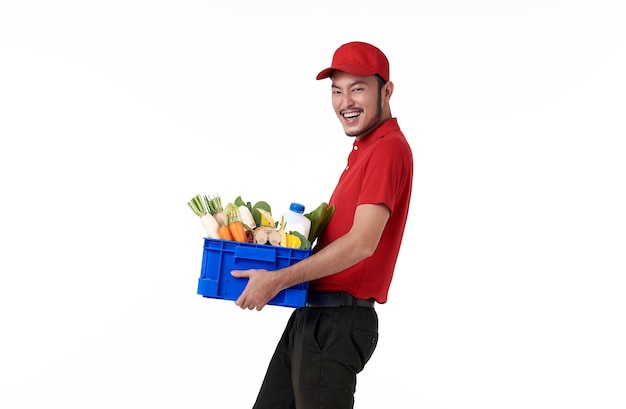 Repartidor asiático vistiendo uniforme rojo con canasta de comida fresca aislada sobre pared blanca.