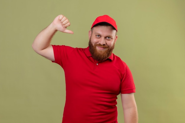 Repartidor barbudo joven complacido en uniforme rojo y gorra apuntando a sí mismo sonriendo confiado