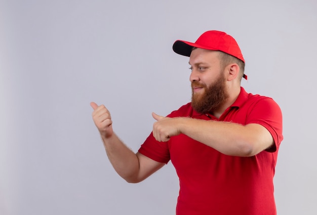Repartidor barbudo joven en uniforme rojo y gorra apuntando hacia atrás con los pulgares
