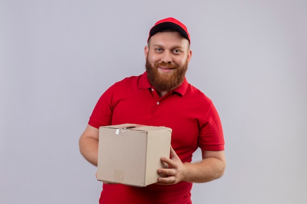 Repartidor barbudo joven en uniforme rojo y gorra con caja de cartón mirando a la cámara con sonrisa de confianza 2