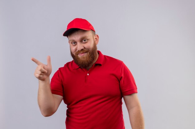 Repartidor barbudo joven en uniforme rojo y gorra mirando confiado apuntando con el dedo hacia el lado