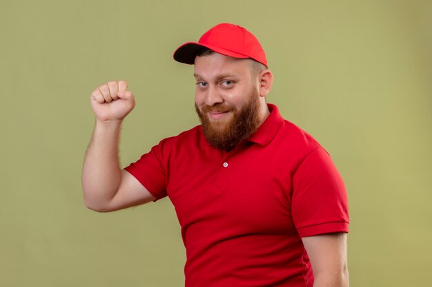 Repartidor barbudo joven en uniforme rojo y gorra sonriendo amigable levantando el puño como ganador, feliz y positivo 2