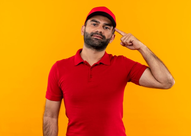 Repartidor barbudo en uniforme rojo y gorra apuntando con el dedo su sien se centró en la tarea de pie sobre la pared naranja
