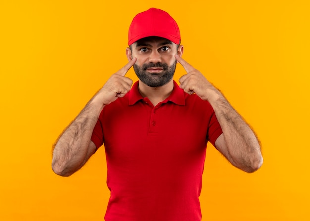 Repartidor barbudo en uniforme rojo y gorra apuntando con los dedos a los ojos de pie sobre la pared naranja