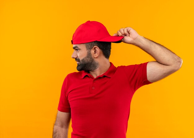 Repartidor barbudo con uniforme rojo y gorra mirando a un lado sonriendo giró su gorra de pie sobre la pared naranja