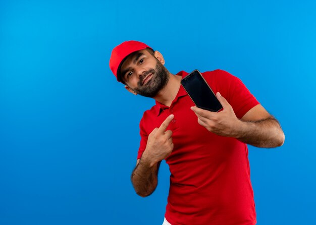 Repartidor barbudo en uniforme rojo y gorra mostrando su smartphone apuntando con el dedo sonriendo de pie sobre la pared azul