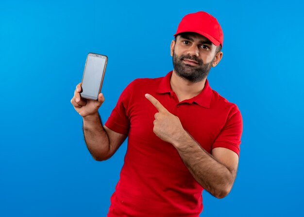 Repartidor barbudo en uniforme rojo y gorra mostrando su teléfono inteligente apuntando con el dedo a él de pie sobre la pared azul