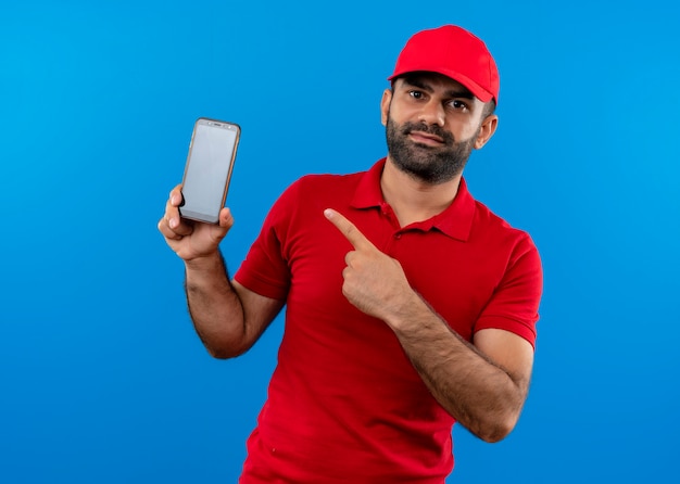 Repartidor barbudo en uniforme rojo y gorra mostrando su teléfono inteligente apuntando con el dedo a él de pie sobre la pared azul