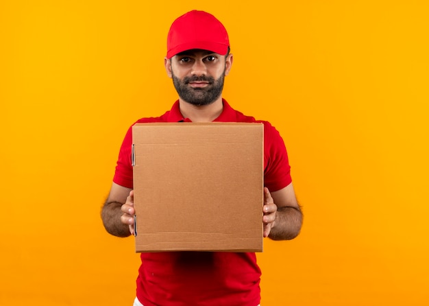 Repartidor barbudo en uniforme rojo y gorra con paquete de caja con expresión seria y segura de pie sobre la pared naranja