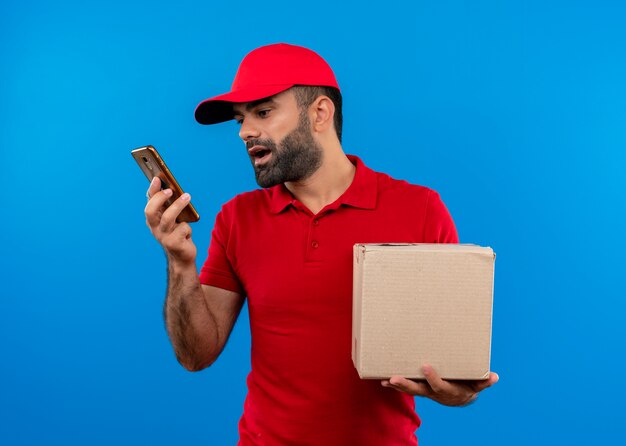 Repartidor barbudo en uniforme rojo y gorra con paquete de caja de grabación de mensaje de voz con su teléfono inteligente de pie sobre la pared azul