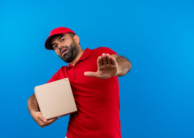 Repartidor barbudo en uniforme rojo y gorra con paquete de caja haciendo señal de stop con expresión de miedo de pie sobre la pared azul