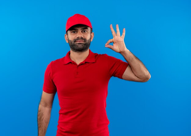 Repartidor barbudo en uniforme rojo y gorra sonriendo confiado mostrando signo ok de pie sobre la pared azul