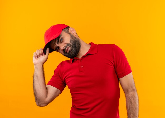 Repartidor barbudo en uniforme rojo y gorra sonriendo y guiñando un ojo de pie sobre la pared naranja