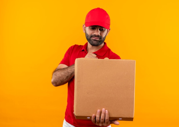 Repartidor barbudo en uniforme rojo y gorra sosteniendo la caja de pizza abierta mirándola sorprendida mirándola con cara seria de pie sobre la pared naranja