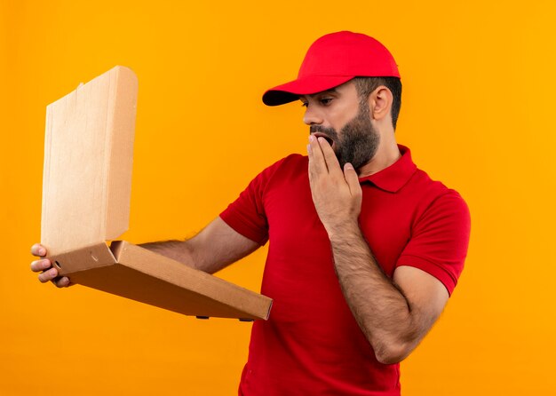 Repartidor barbudo en uniforme rojo y gorra sosteniendo una caja de pizza abierta mirándola sorprendida de pie sobre la pared naranja