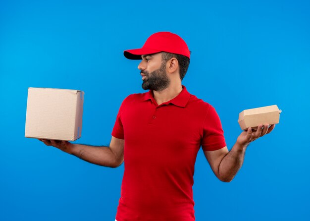 Repartidor barbudo en uniforme rojo y gorra sosteniendo cajas de cartón con aspecto confundido teniendo dudas de pie sobre la pared azul